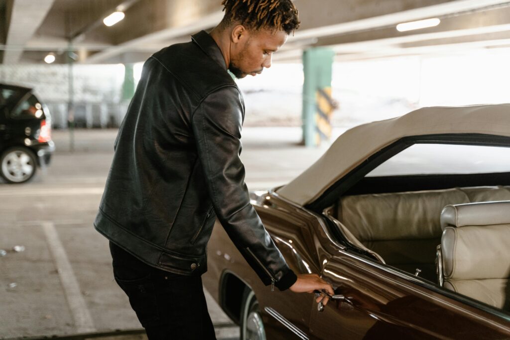 African American man in black leather jacket opening a door of a vintage car in a parking area.