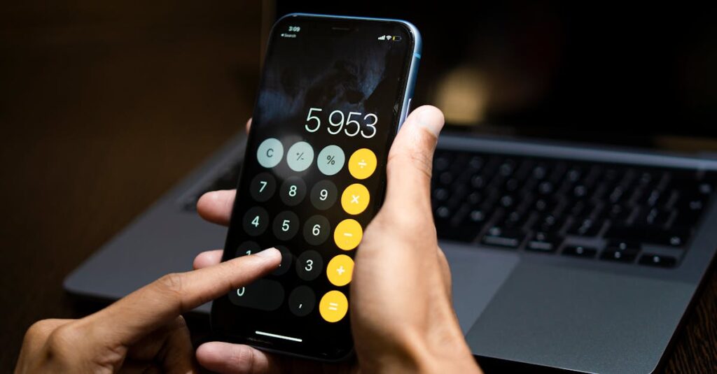 Person using a calculator app on a smartphone with a laptop in the background.